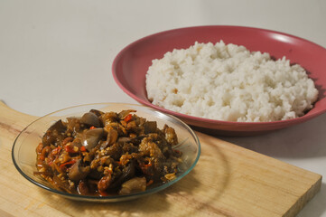 Stir-fried purple eggplant with anchovies and peanuts served in a small plate on a wooden cutting board and a plate of white rice isolated on a white background