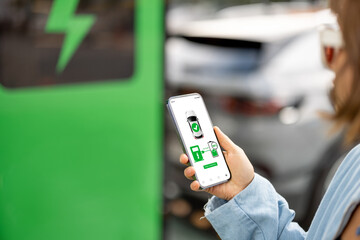Woman holds a smartphone with running application for vehicle charging, electric car charging on background, close-up