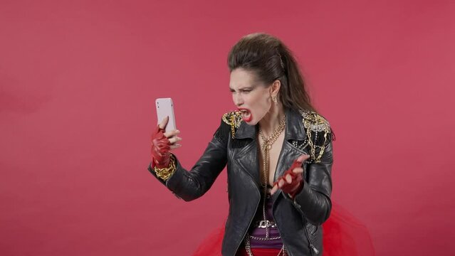A woman makes a video call on a smartphone, speaks aggressively, ends the conversation and crosses her arms. Angry, upset woman in the studio on a red background. Advertising. HDR BT2020 HLG Material.
