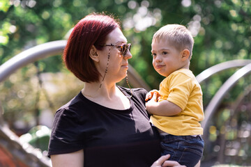 Loving mother in sunglasses looking at little son with Down syndrome in arms woman and cute boy spending time together at city playground on sunny summer day