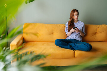 Portrait of pensive stressed young woman sitting on sofa at home using smartphone looking away pondering of problem. Anxious redhead female feel distressed frustrated with message or text on phone.