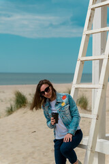 a girl with a glass of coffee stands on the beach near the rescue tower