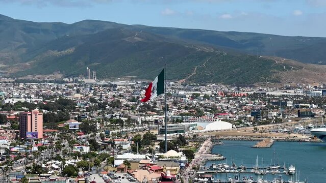 The flag of Mexico in the central zone of Ensenada against the backdrop of a city landscape and mountains. The flag flutters in the wind, slow motion aerial view. 