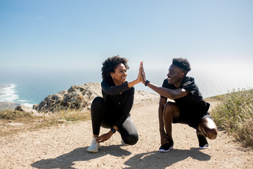 Excited young black couple tying shoelaces, giving high five, enjoy workout together in morning on coast landscape