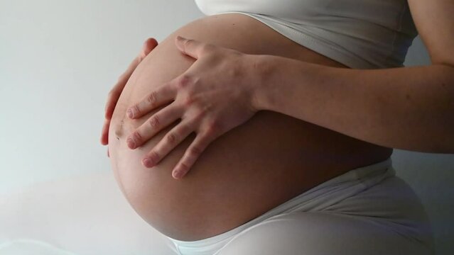 Midsection of heavily pregnant baby belly held by hands of woman bouncing on gym ball. Side view. White background. Bright shot.