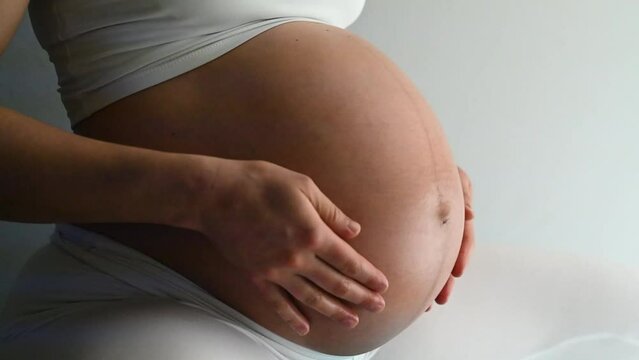 Midsection of heavily pregnant baby belly held by hands of woman bouncing on gym ball. Side view. White background. Bright shot.