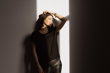 Smiling young female in casual clothes touching hair and looking away camera while leaning on white wall in light room