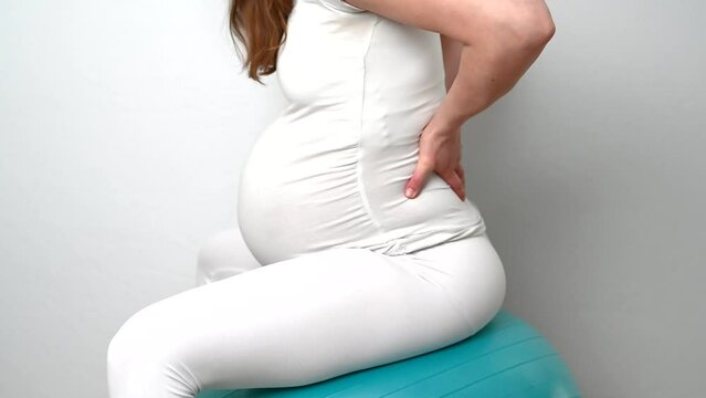 Midsection of heavily pregnant baby bump held by hands of woman bouncing on exercise ball. Side view. White background. Bright shot.