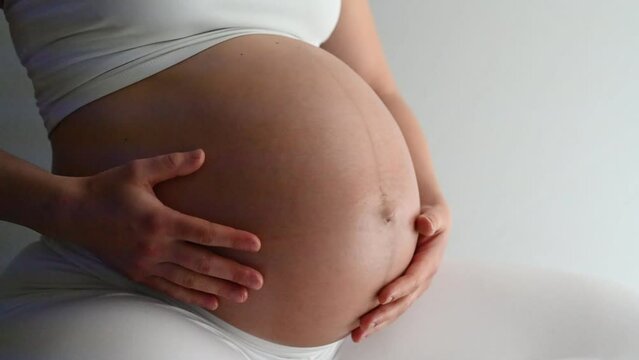 Midsection of heavily pregnant baby bump held by hands of woman bouncing on exercise ball. Side view. White background. Bright shot.