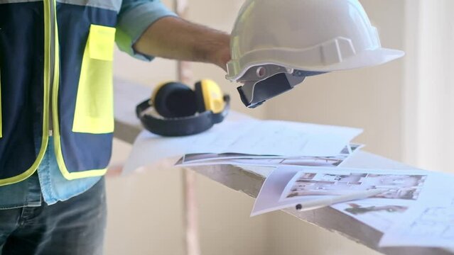 Architect in vest taking white helmet from heap of project papers to continue renovation process in bedroom man in uniform at construction site safety rules and equipment