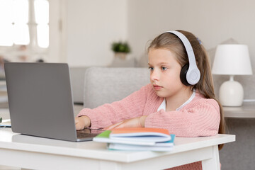 Serious millennial caucasian teenager girl in headphones studying, doing homework, watching online lesson