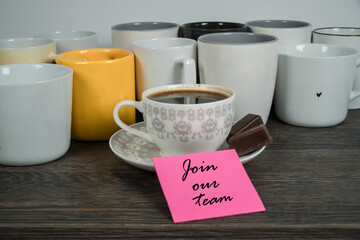 Kawa w filiżance z czekoladkami i kartką z napisem: Join our team. Filiżanka otoczona innymi kubkami. Przywitanie w zespole. - obrazy, fototapety, plakaty