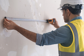 Focused engineer in protective helmet measuring shabby wall with tape-line preparing for drilling holes man in uniform helping owners to do renovation process in apartment