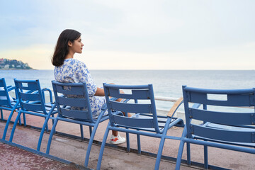 Beautiful smiling young mixed race woman sitting at one of the famous blue chairs at the Promenade des Anglais in Nice, France