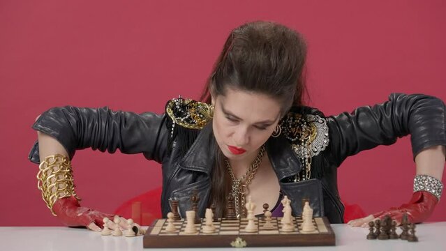 Angry woman sits at a table with chessin the studio on a red background. The woman throws off all the pieces from the chessboard with her hand, gets up and leaves. HDR BT2020 HLG Material.