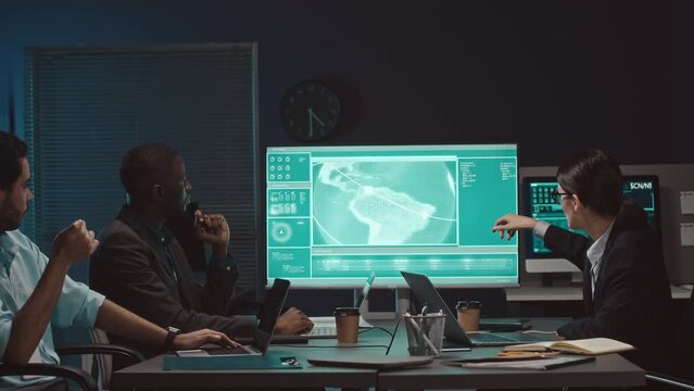 Three multiethnic cybersecurity workers looking at round globe world map on computer monitor and making analysis, working together in dark cyber security agency at night