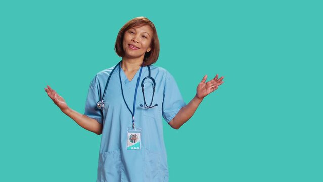 Puzzled asian nurse doing perplexed gesturing while at work. Clueless confused healthcare expert at a loss of words over what she hears, isolated over blue studio background
