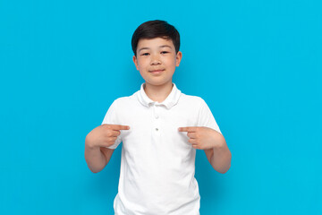 little asian boy in white t-shirt points to himself on blue background, korean child advertises copy space