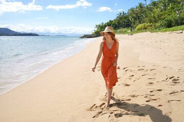 Young woman with orange sundress and straw hat walking on the sand beach - obrazy, fototapety, plakaty