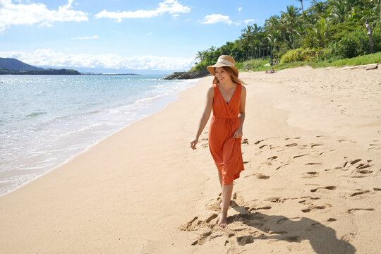 Young woman with orange sundress and straw hat walking on the sand beach