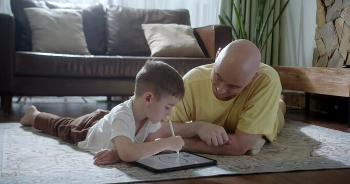 Happy family, young parents lying on the warm floor at home, dad and son homeschooling on a tablet device in the living room at leisure. 4K