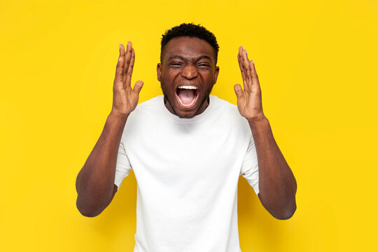 aggressive african american man in white t-shirt screams from headache and fatigue on yellow background