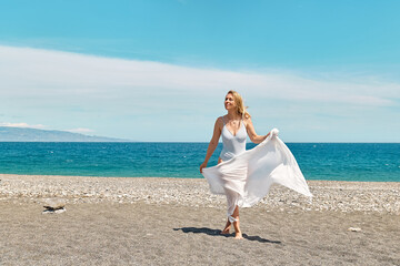 Fototapeta na wymiar Pretty blond woman in swimsuit and white pareo enjoying sunny windy day near blue sea. Happy female enjoying the life. Beach summer, travel concept.