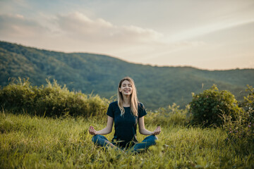 The rising sun casts a warm glow on a woman as she peacefully sits in a lotus position, finding tranquility in nature.