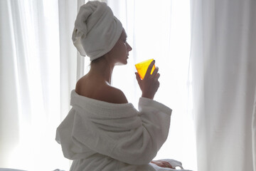 Young beautiful woman wrapped in towels drinking liquid collagen or vitamin supplement next to the...