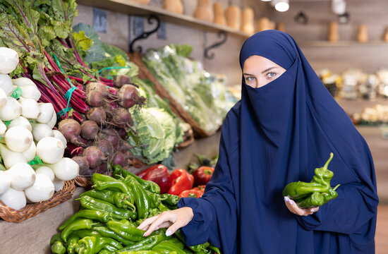 Portrait of girl in paranja buying fresh bell peppers in grocery shop