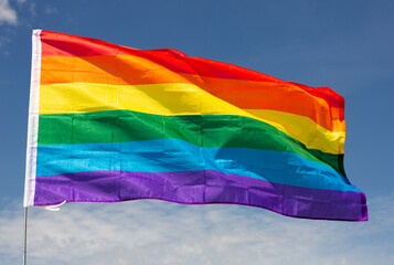 Large LGBT flag fixed on metal stick waving against background of clear sky during daytime