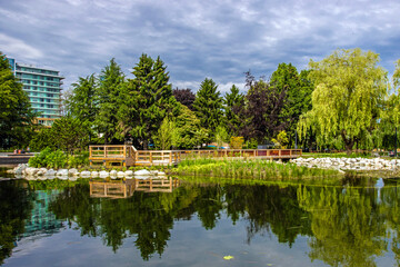 Fototapeta na wymiar Beautiful summer day, a pond in a city park, a wooden bridge and a promenade over the water, a green tree and grass, buildings in the background and a dramatic cloudy sky