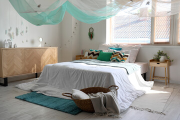 Interior of modern bedroom with bed, chest of drawers and dream catcher hanging on white wall