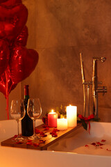 Wooden tray with wine, burning candles and rose petals on tub in bathroom. Valentine's day celebration