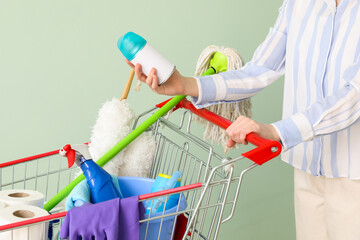 Woman with shopping cart full of cleaning supplies on pale green background