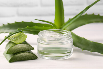 Jar of cosmetic gel and cut aloe vera leaves on white wooden table, closeup