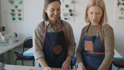 Medium close-up tilting shot of middle-aged female leather atelier owner and young apprentice standing together by cutting table and choosing leather for new products