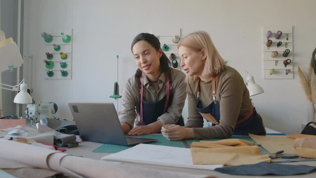 Medium shot of two female tailors standing in atelier, leaning on cutting table among rolls of leather and suede, watching fashion show on laptop and discussing new models and trends