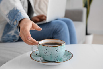 Woman on sofa using laptop and taking cup of coffee from table, closeup