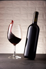 Dynamic image of unlabeled red wine bottle and wine glass with wine flowing into it. Gray stone surface, white rustic brick wall background. minimalist concept.