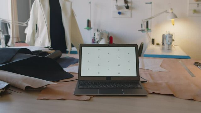 Full arc shot of laptop with empty white screen on table of fashion designer in leather atelier, unfinished jacket on tailors dummy and professional sewing machines in background. Template, copy space