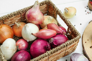 Wicker basket with different kinds of onion on white wooden background