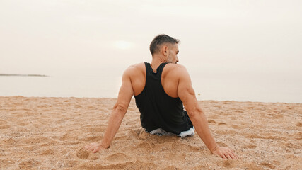 Fototapeta na wymiar Middle-aged athletic man sits on the beach after workout and looks at the sea, Back view