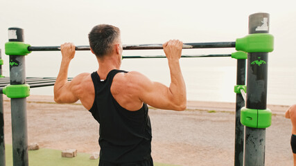 Naklejka premium Middle-aged muscular man in sportswear pulls himself up on the horizontal bar on sea background, Back view