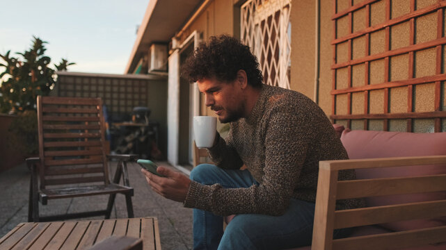Happy guy sitting on the terrace drinks coffee, browses the web on smartphone - Powered by Adobe