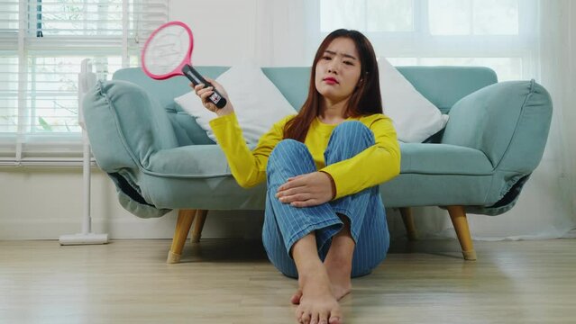 Asian young woman sitting floor using mosquito swatter or electric net racket, female killing mosquitoes she holding fly swatter like weapon in living room at home, Insect killer