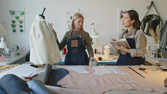Medium full shot of middle-aged experienced female tailor showing work process of sewing leather jacket to young apprentice, who is listening and writing in notebook