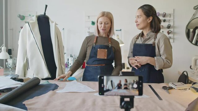 Selective focus zoom out shot of smartphone on tripod, and two female tailors in aprons standing in front of camera, next to dummy, talking and recording sewing tutorial for online course