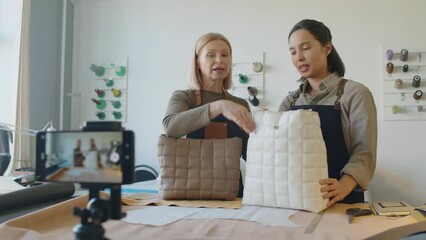 Medium shot of leather workshop owner and employee filming story or reel on smartphone for social media about new handbag, and demonstrating its features for subscribers