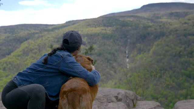 Young diverse woman hiker pets her brown senior dog at hiking trail viewpoint rocky cliff overlooking a beautiful picture perfect view of a forest mountainside waterfall 4K SLOW MOTION depth of field